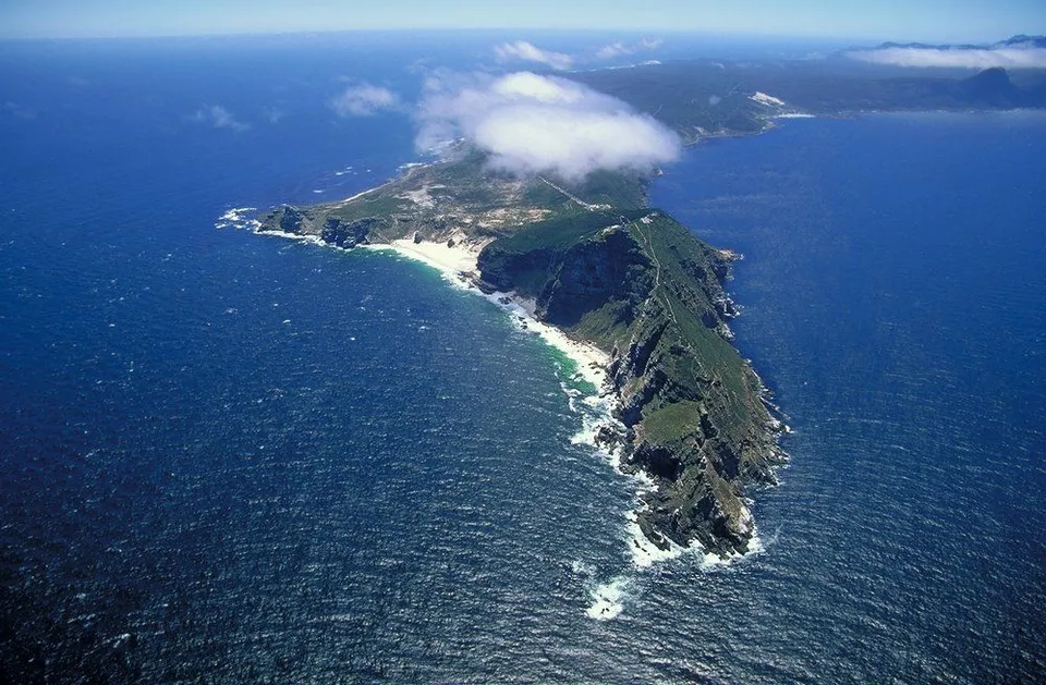 Aerial view of the rugged, green Cape Peninsula coastline jutting into the deep blue ocean, with white sand beaches and clouds overhead.