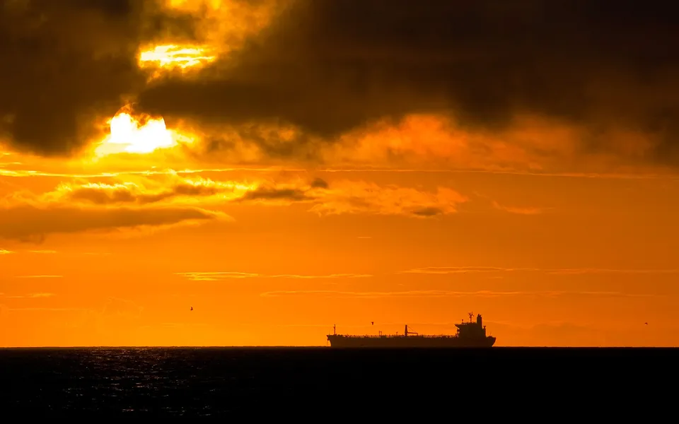 A large cargo ship is silhouetted against a vibrant orange sunset sky over the dark ocean.