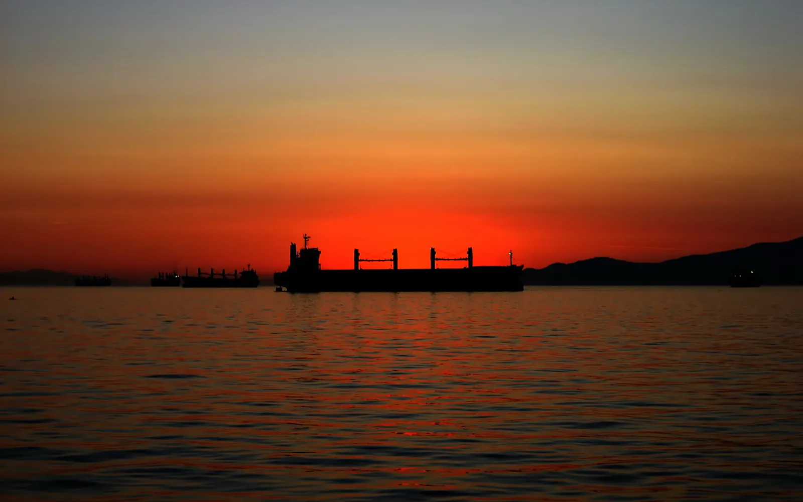 Silhouetted cargo ships on the water with a vibrant orange and red sunset sky.