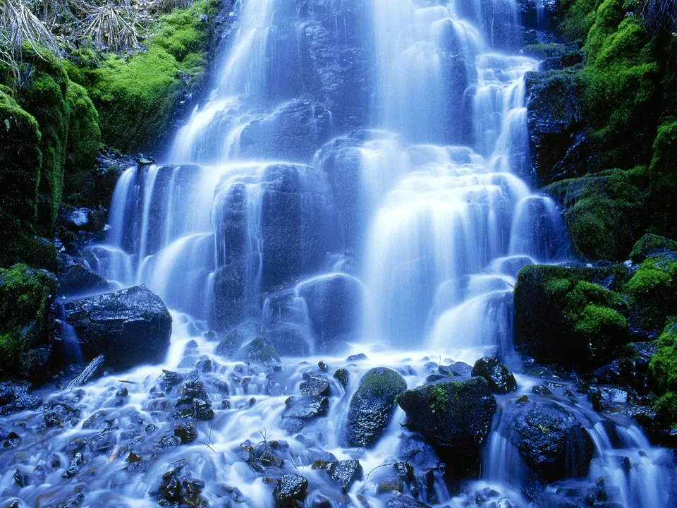 A multi-tiered waterfall flows over moss-covered rocks in a lush forest.