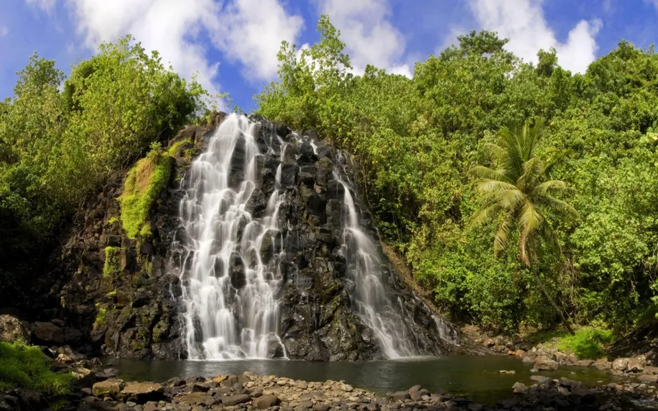 A wide tropical waterfall cascades down a dark, jagged rock face surrounded by lush green foliage under a blue sky.