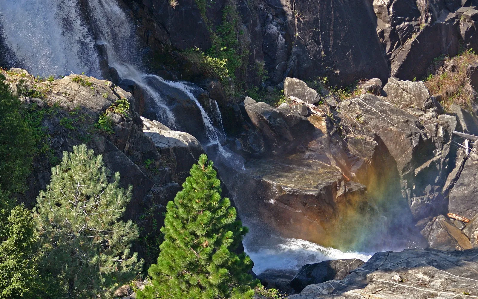 Sunlight creates a rainbow in the mist of a cascading waterfall framed by trees.