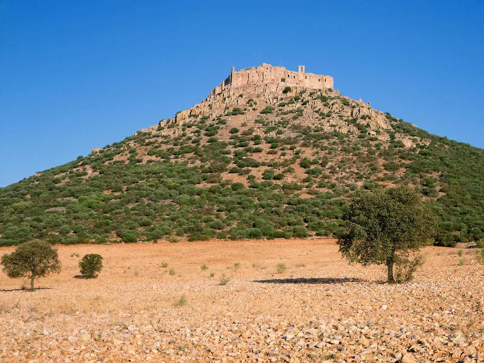 A stone castle sits atop a hill covered in green shrubs, with a rocky, dry foreground and a clear blue sky.
