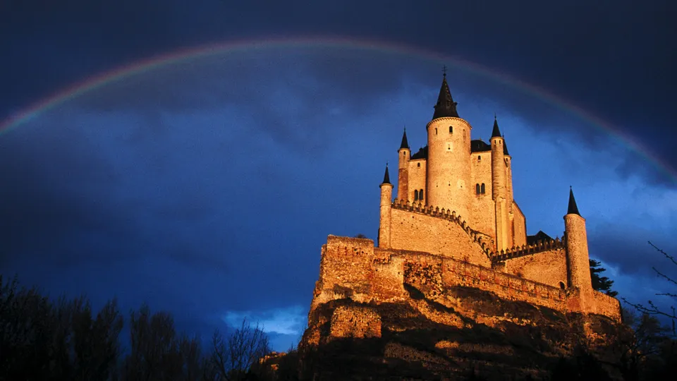 A medieval castle perched on a hill under a dark blue sky with a rainbow arching over it.