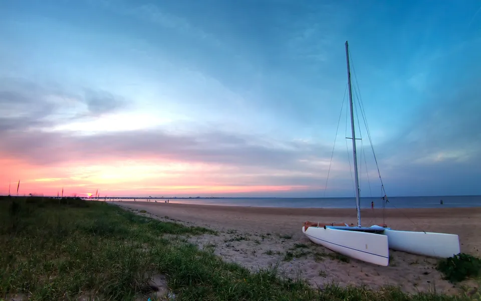A white catamaran rests on a sandy beach with the ocean and a colorful sunset sky in the background.
