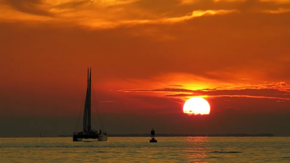 A catamaran sails on the water as the sun sets, casting a fiery orange glow across the sky and sea.