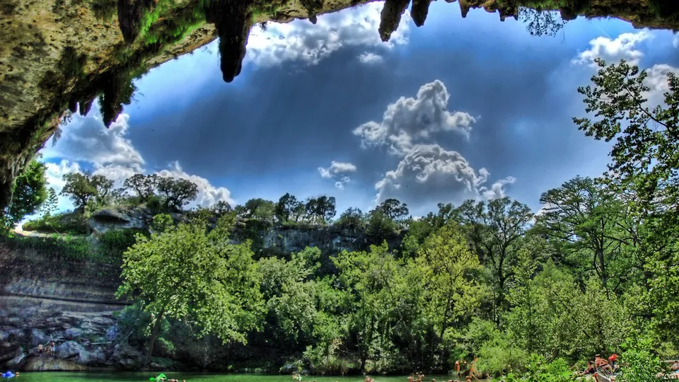 View from inside a cave looking out at a green river, lush trees, and a blue sky with clouds.