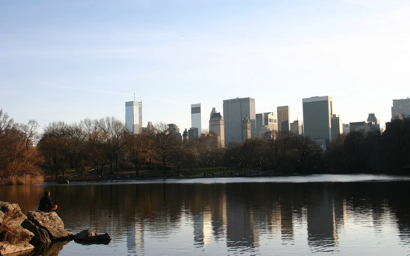 A person sits on rocks by a lake, with a city skyline reflected in the water.