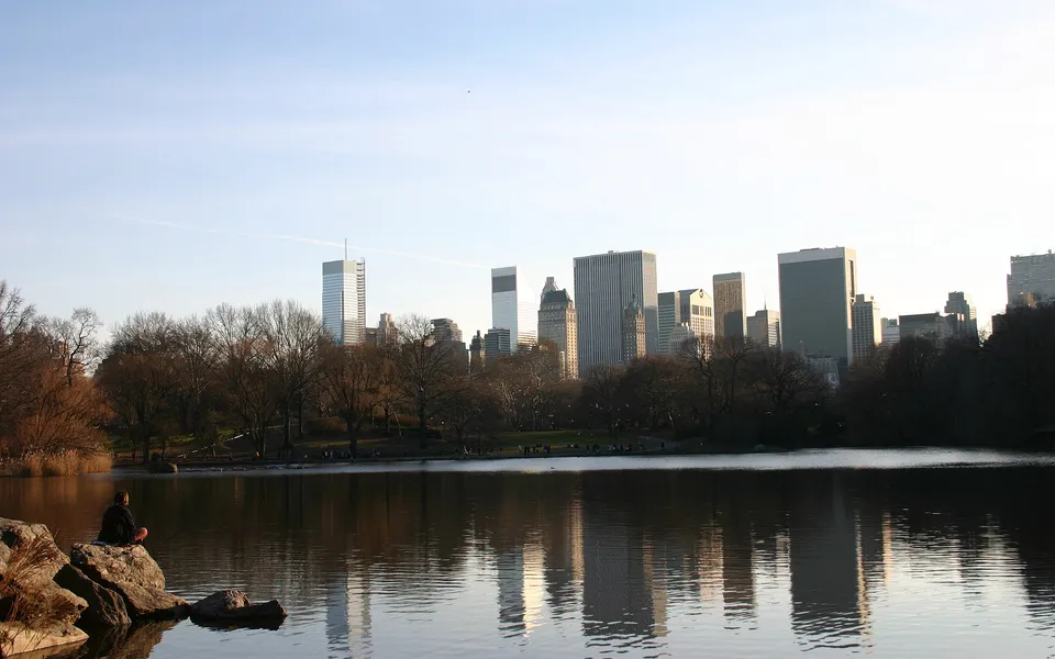 A person sits on rocks by a lake, with a city skyline reflected in the water.
