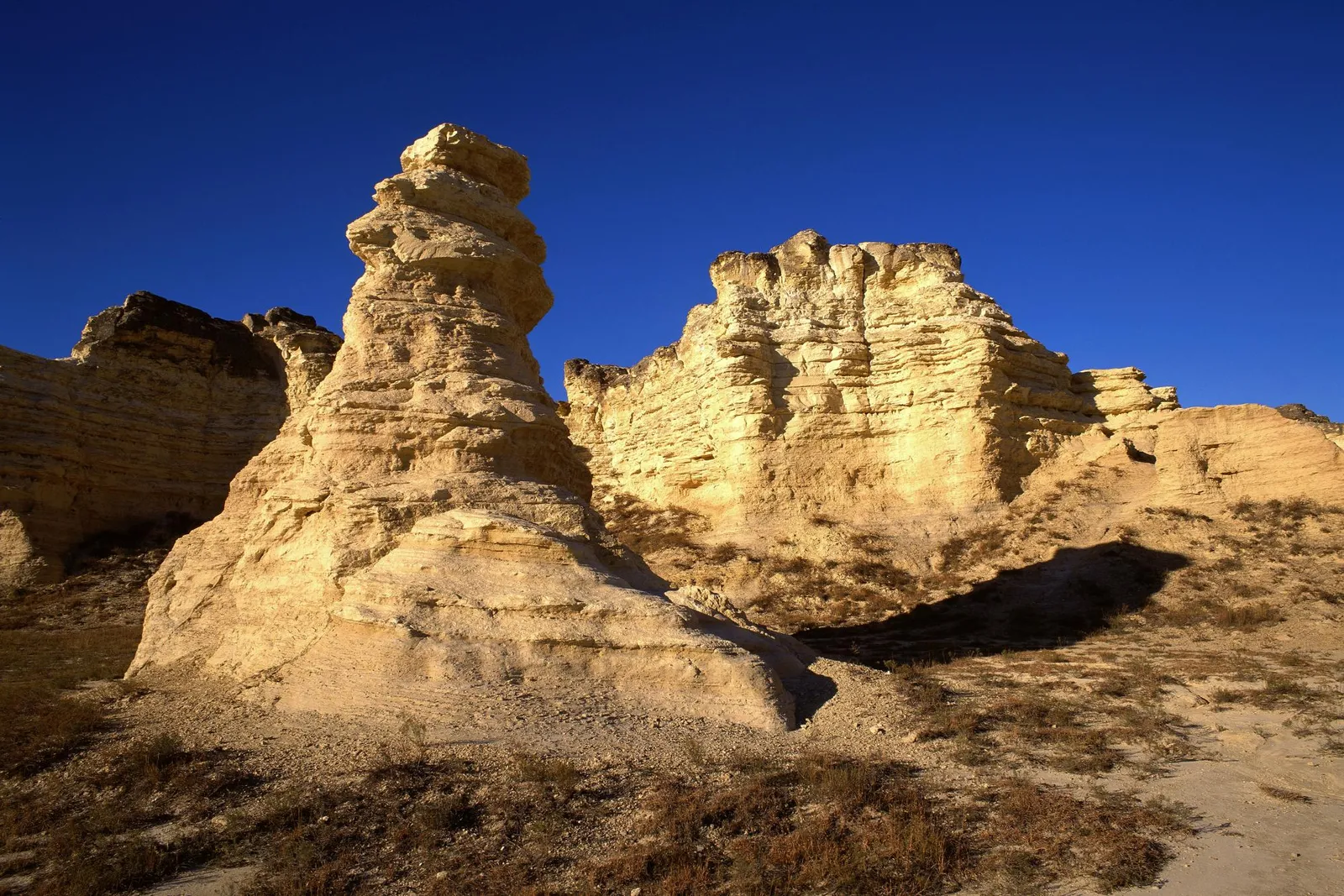 Tall, layered chalk rock formations rise from a dry, grassy landscape under a deep blue sky.