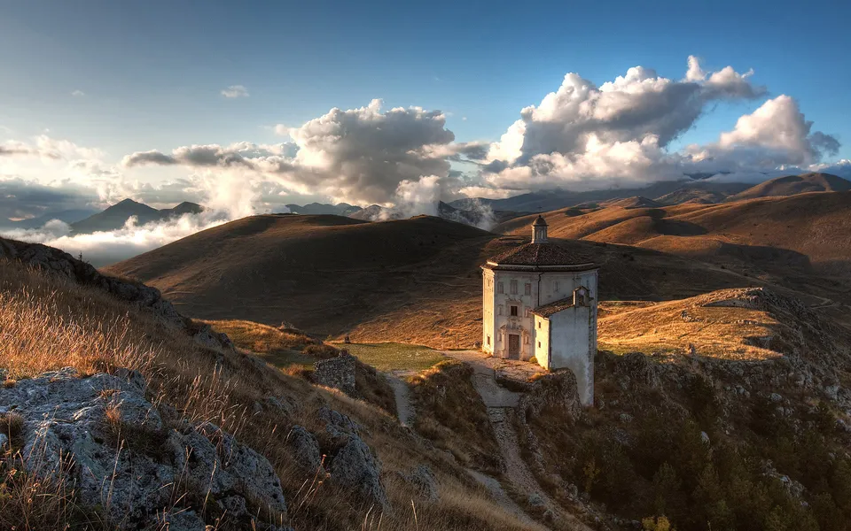A white chapel sits on a hilltop with rolling hills and dramatic clouds in the background during golden hour.