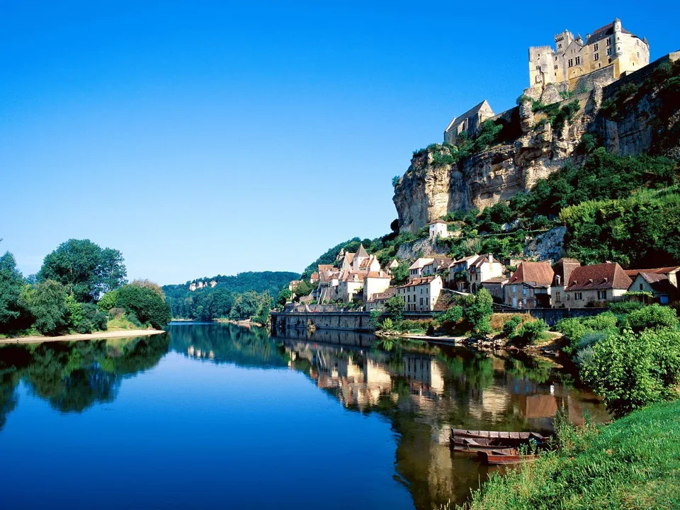 A medieval castle and village sit atop a cliff overlooking a calm river with reflections.