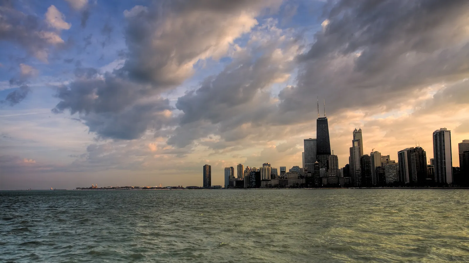 A wide view of the Chicago skyline with tall buildings silhouetted against a dramatic sky with clouds.