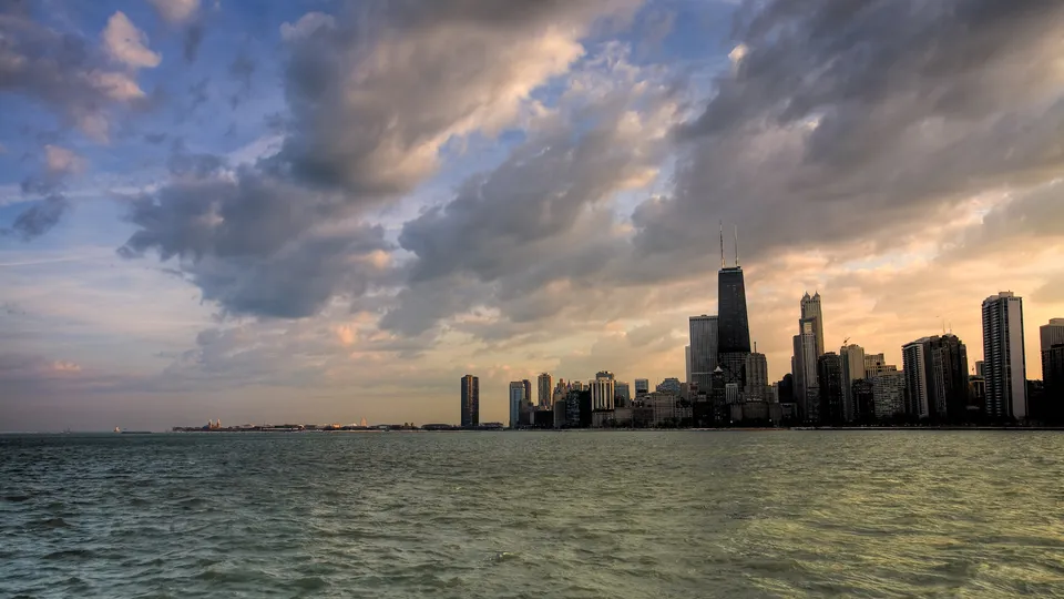 A wide view of the Chicago skyline with tall buildings silhouetted against a dramatic sky with clouds.