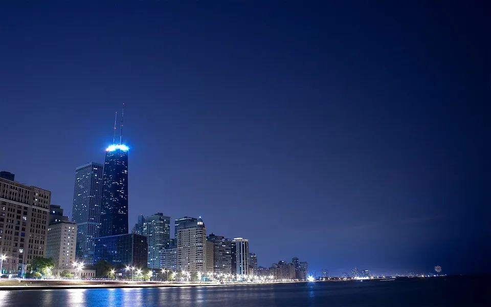 A wide shot of the Chicago skyline at night, with illuminated skyscrapers reflecting on the water.