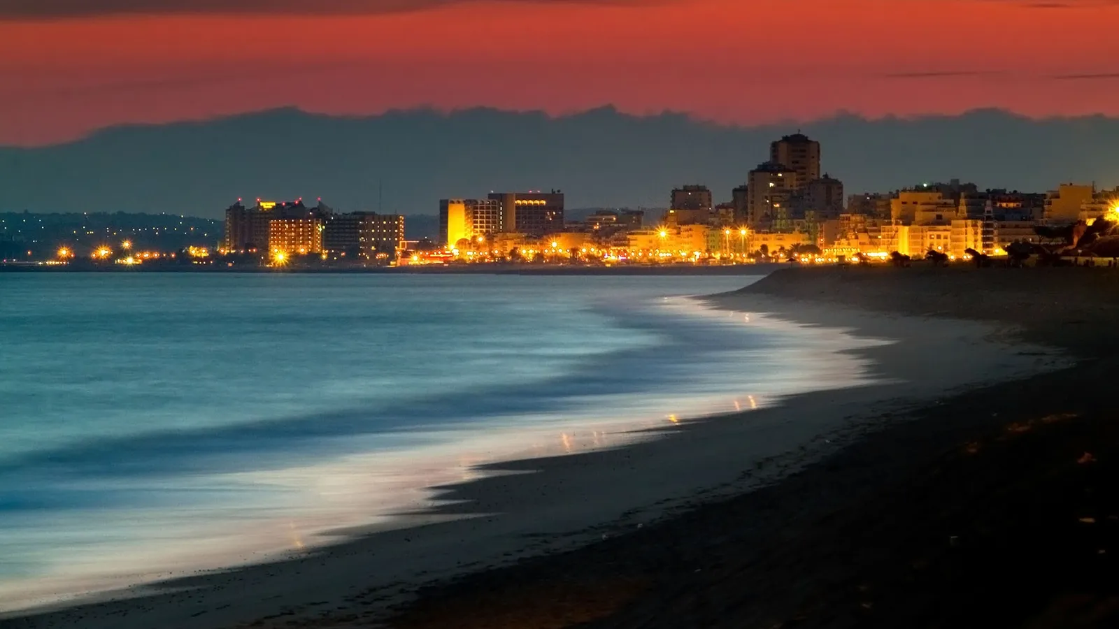 A beach with gentle waves lapping the shore, leading to a city skyline illuminated by lights under a colorful twilight sky.