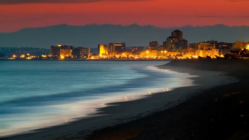 A beach with gentle waves lapping the shore, leading to a city skyline illuminated by lights under a colorful twilight sky.