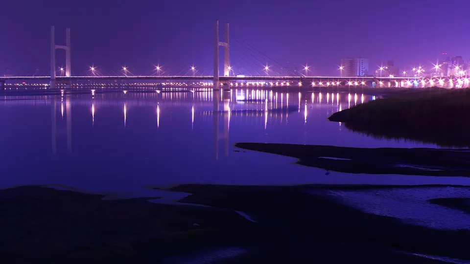 A long suspension bridge with lights is reflected in the dark water below, under a purple night sky.