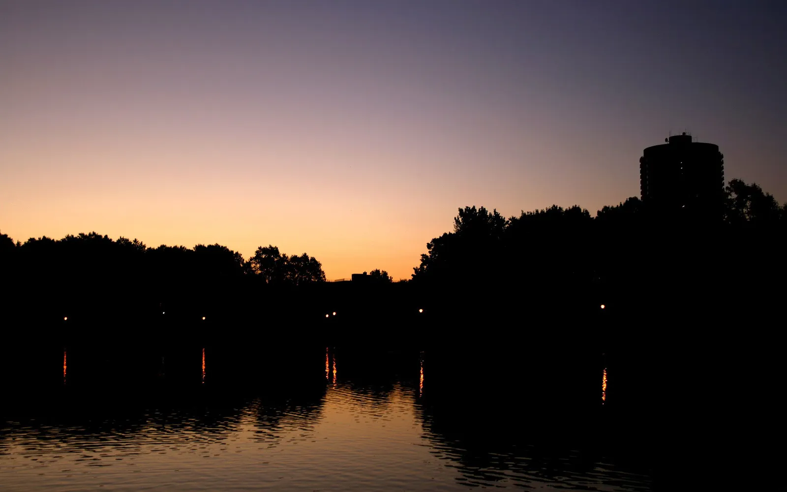 A silhouetted cityscape and trees are reflected in a calm lake during a purple and orange sunset.