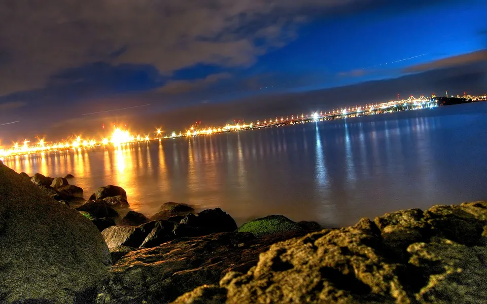 A long exposure shot of city lights reflecting on the water at night, with rocks in the foreground.