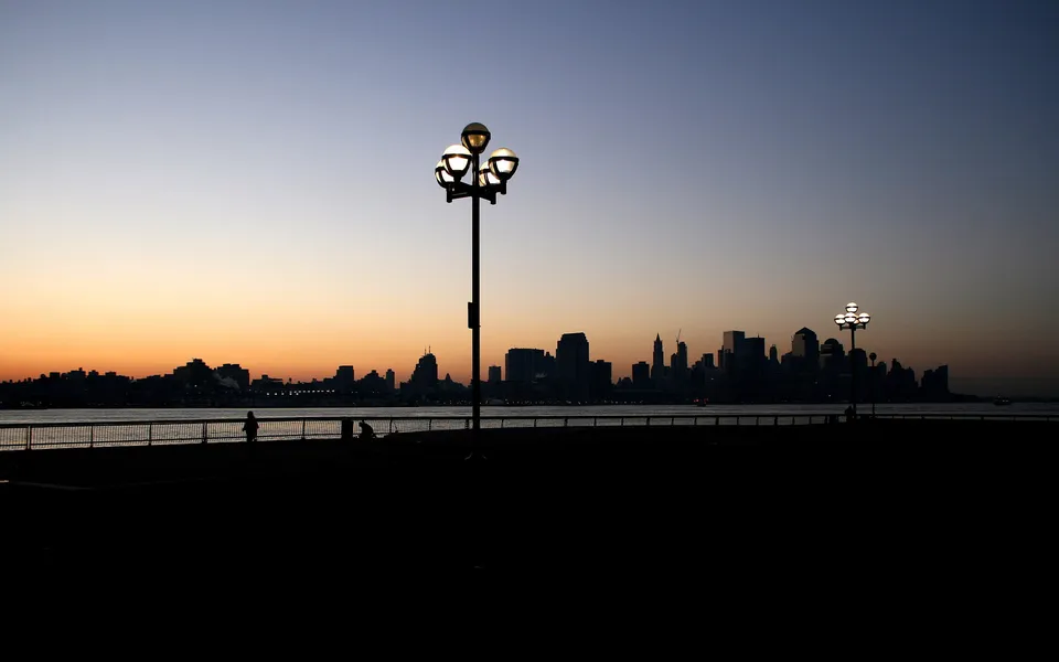 A city skyline is silhouetted against a gradient sky of orange and blue, with lampposts in the foreground.