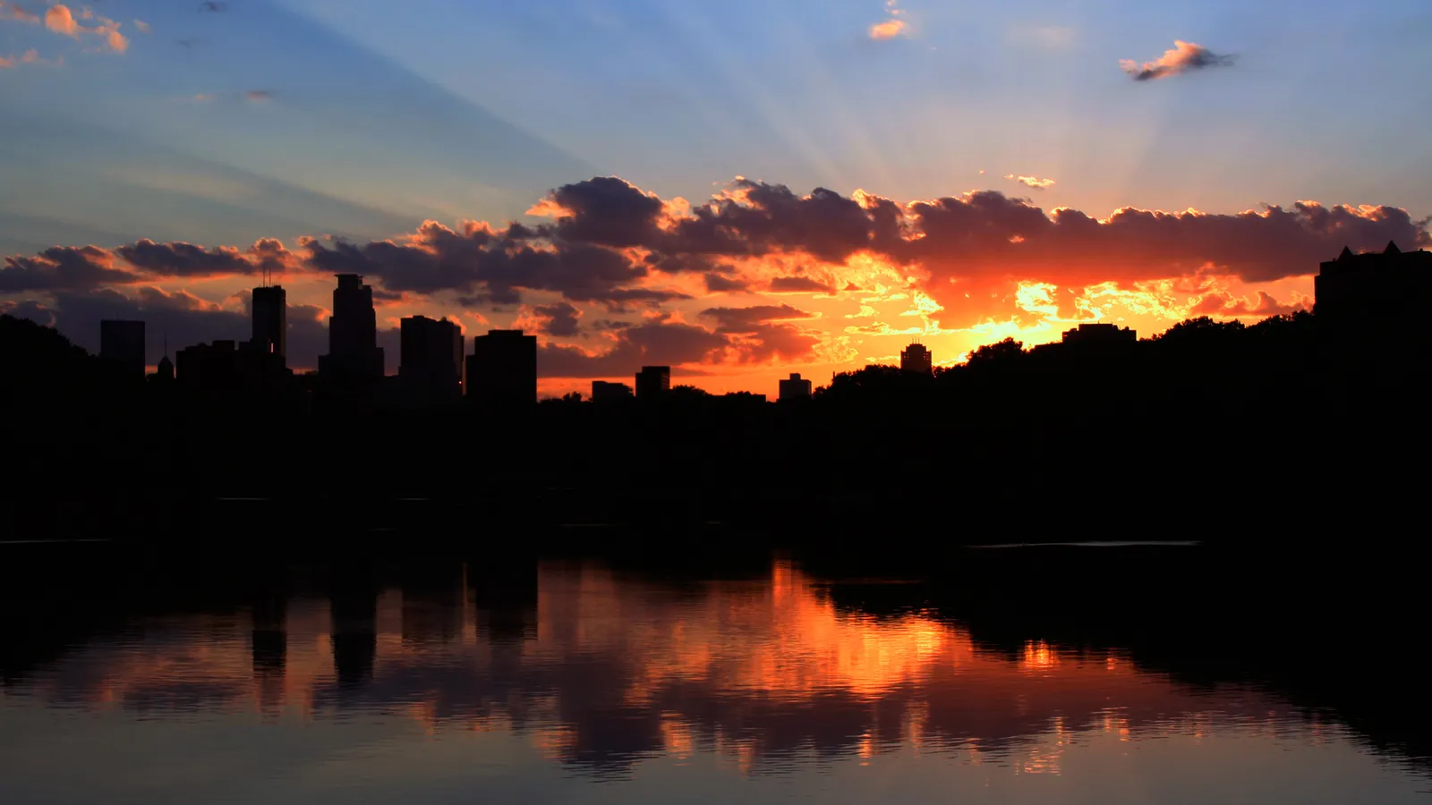 A city skyline is silhouetted against a vibrant sunset sky with crepuscular rays, reflected in the calm water below.
