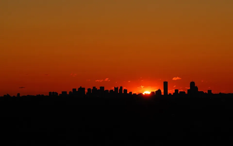 A city skyline is silhouetted against a vibrant orange and red sunset sky.