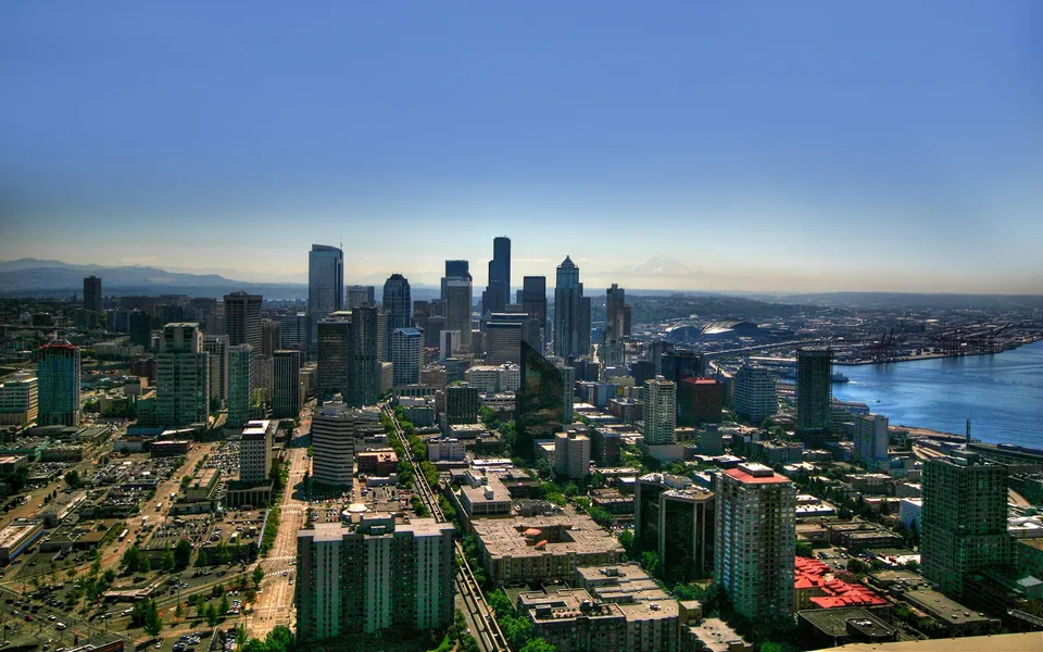 An aerial view of a dense cityscape with tall buildings, a busy harbor, and distant mountains under a clear blue sky.