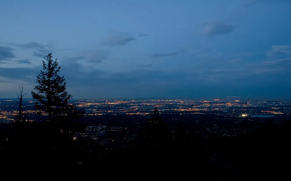 A wide view of a city at dusk, with lights twinkling in the distance, seen from a dark, tree-lined hill.