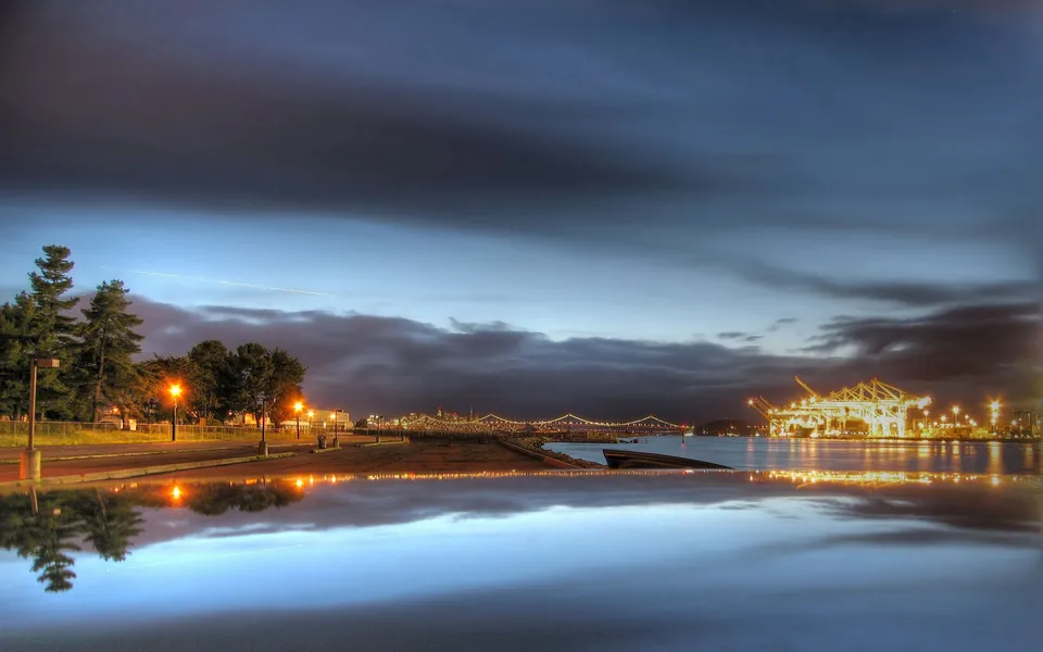 A city skyline with illuminated cranes and a bridge is reflected in calm water under a cloudy sky at dusk.
