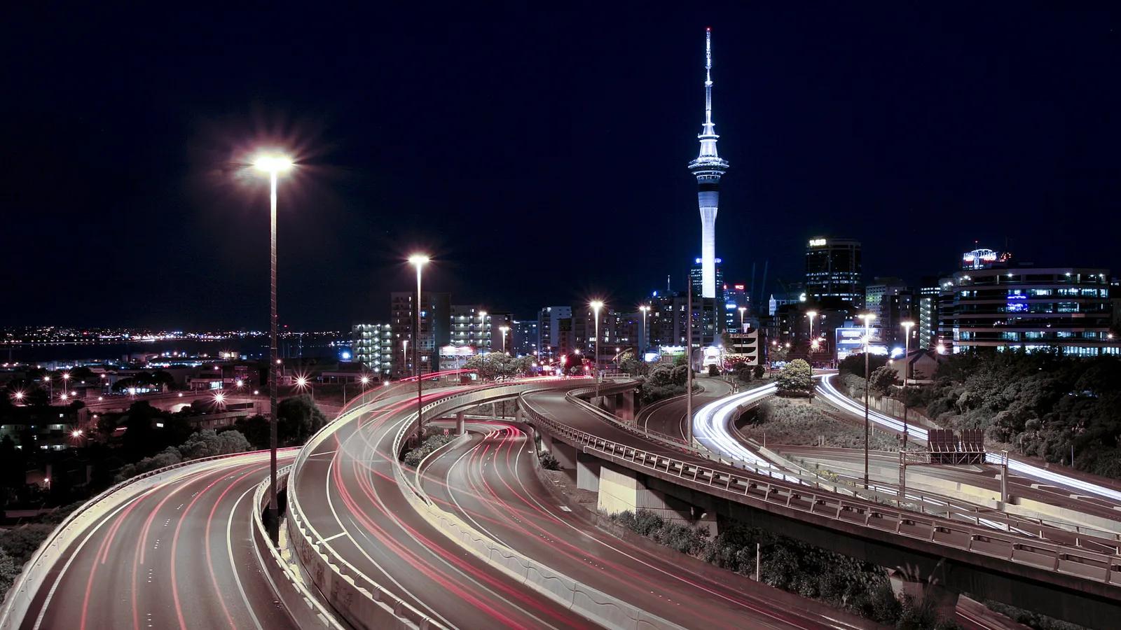 A nighttime cityscape featuring the Sky Tower, illuminated buildings, and light trails from cars on a highway.