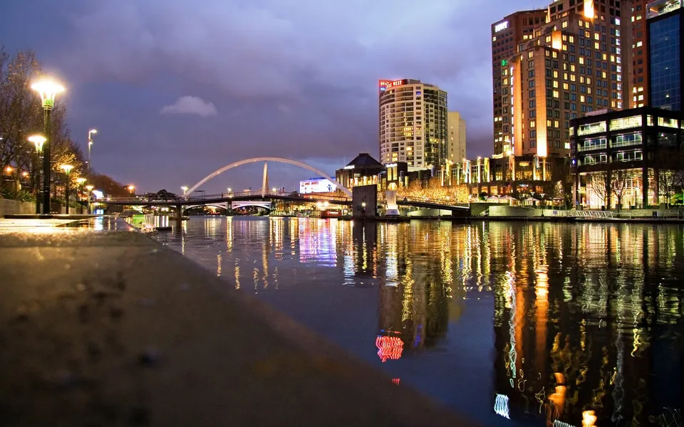 A city skyline with illuminated buildings and bridges reflected in a dark river at dusk.