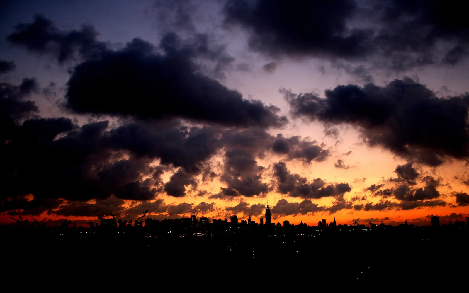 A dark, cloudy sky with vibrant orange and purple hues above a silhouetted city skyline at dusk.