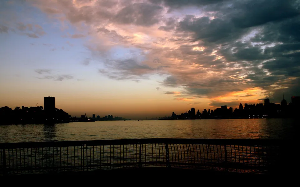 A silhouetted city skyline is visible across a body of water during a colorful sunset with dramatic clouds.