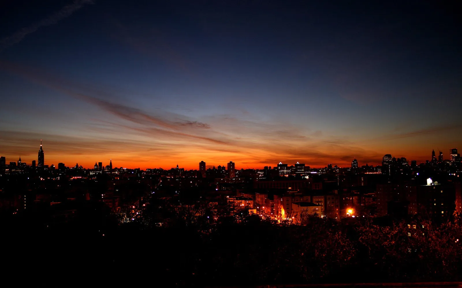 A city skyline is silhouetted against a vibrant orange and deep blue sunset sky with wispy clouds.
