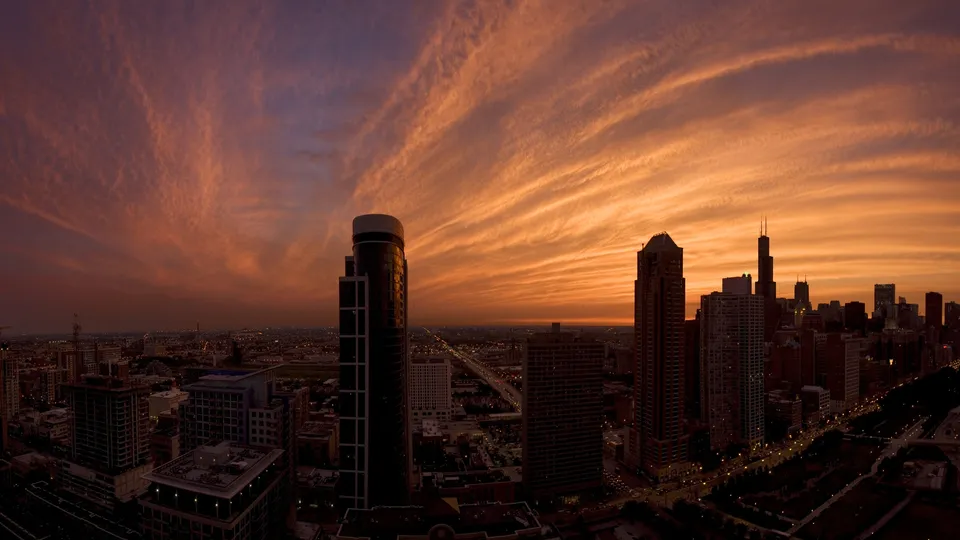 A panoramic view of a city skyline at sunset, with vibrant orange and purple clouds stretching across the sky.