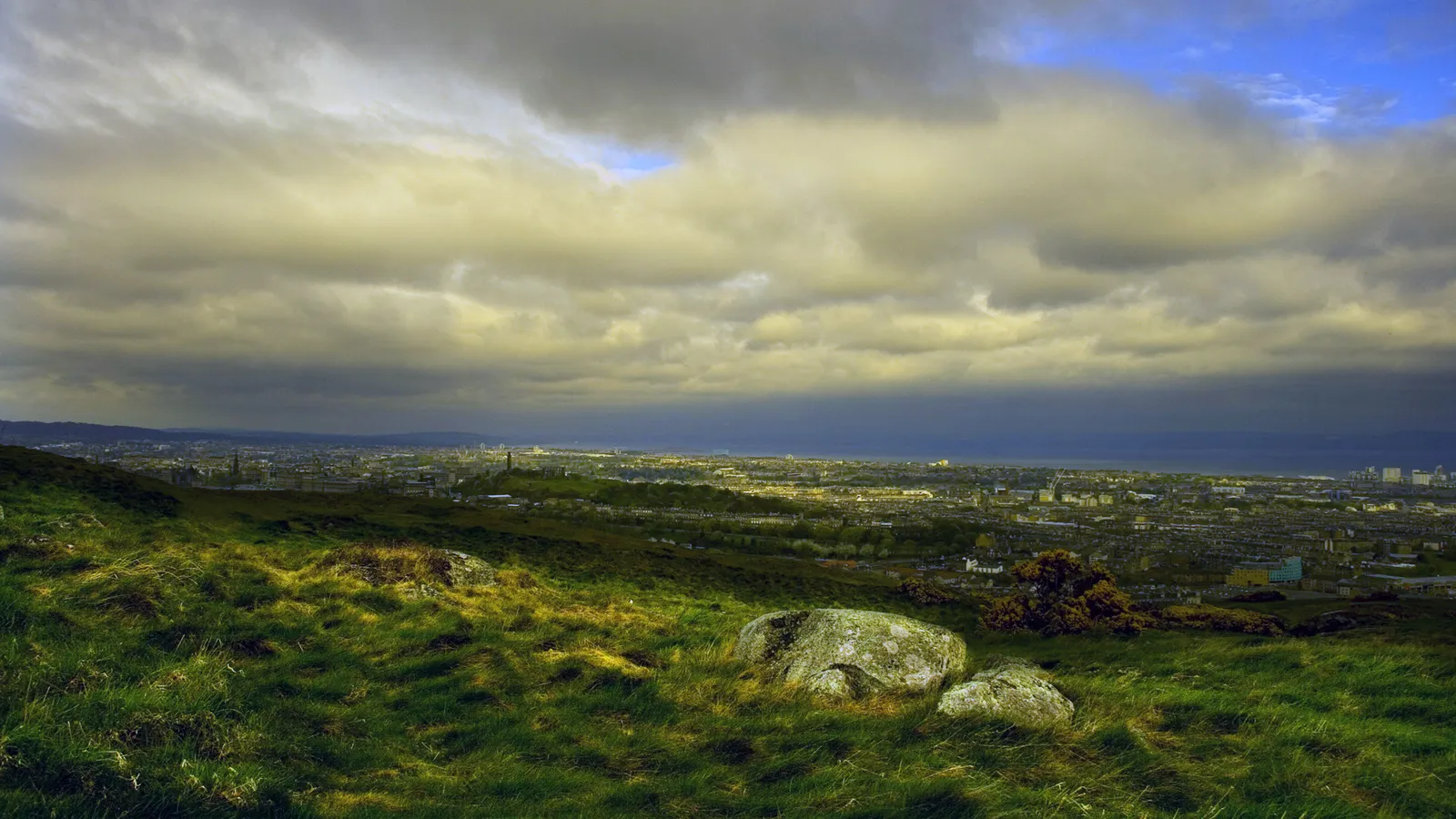 A wide view of a city spread out below a grassy hill with large rocks in the foreground and a dramatic sky above.