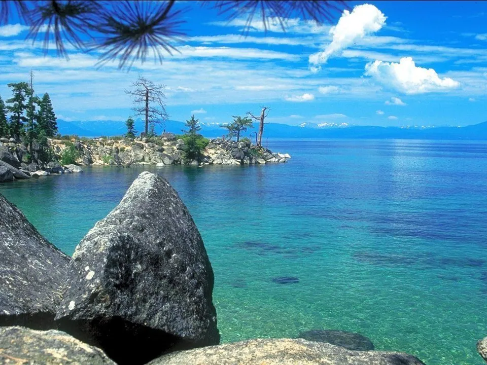 Large grey rocks in the foreground with a clear blue lake and distant mountains under a cloudy blue sky.