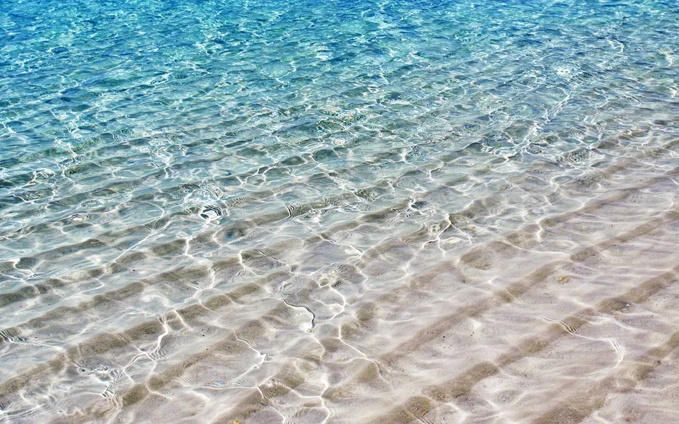 Close-up of shallow, clear turquoise ocean water with sunlight reflecting on the sandy seabed below.