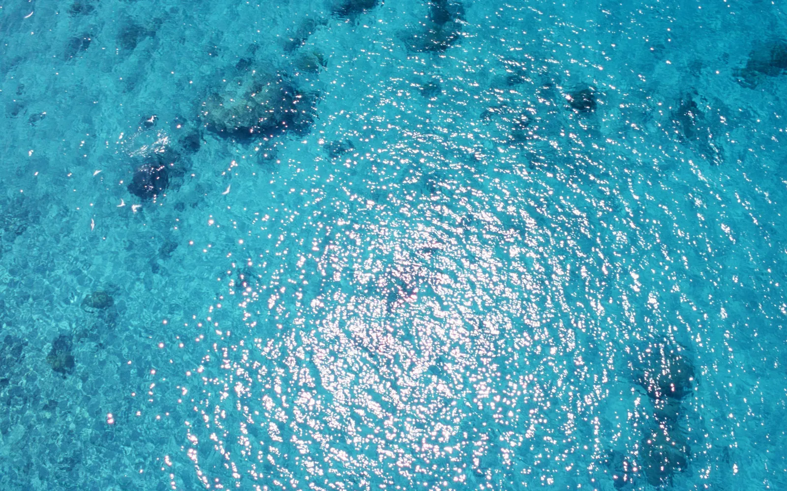 Aerial view of clear turquoise ocean water with sunlight glinting on the surface and coral formations visible below.