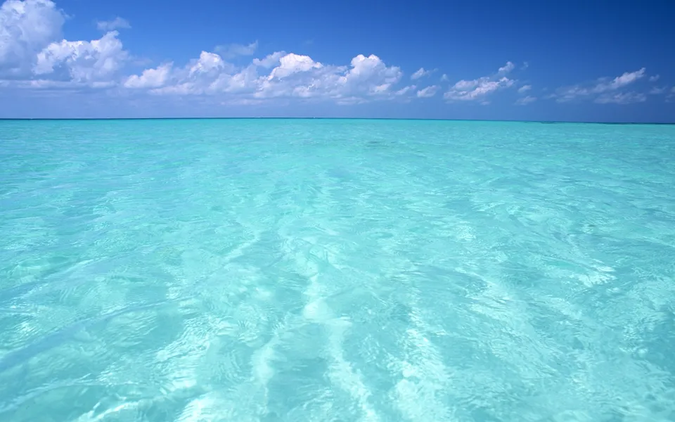 Vast expanse of clear turquoise ocean water with gentle ripples under a bright blue sky with scattered white clouds.