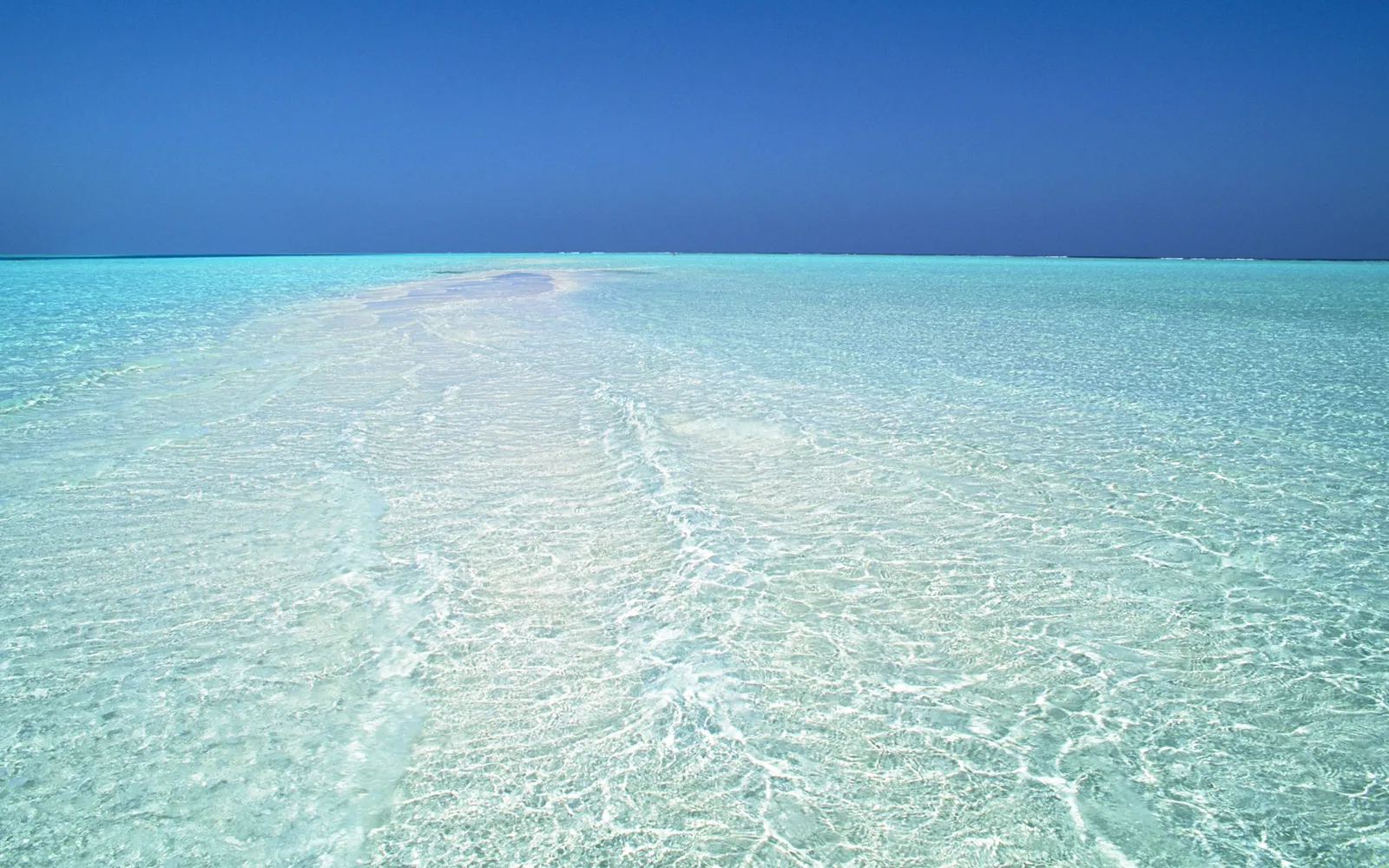 A vast expanse of shallow, crystal-clear turquoise water stretches to the horizon under a deep blue sky.