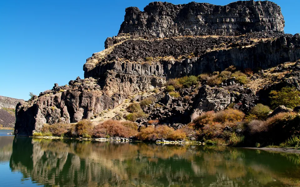 A large, dark, rocky cliff face is reflected in the calm, green water of a lake below.