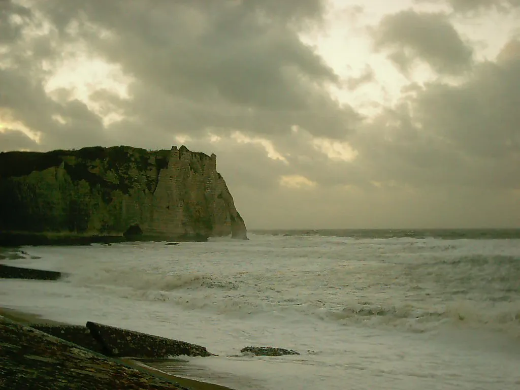 A large, green cliff face rises from a turbulent sea under a cloudy sky.