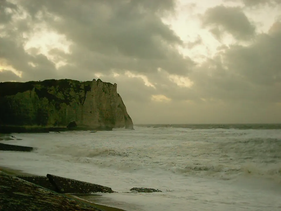 A large, green cliff face rises from a turbulent sea under a cloudy sky.