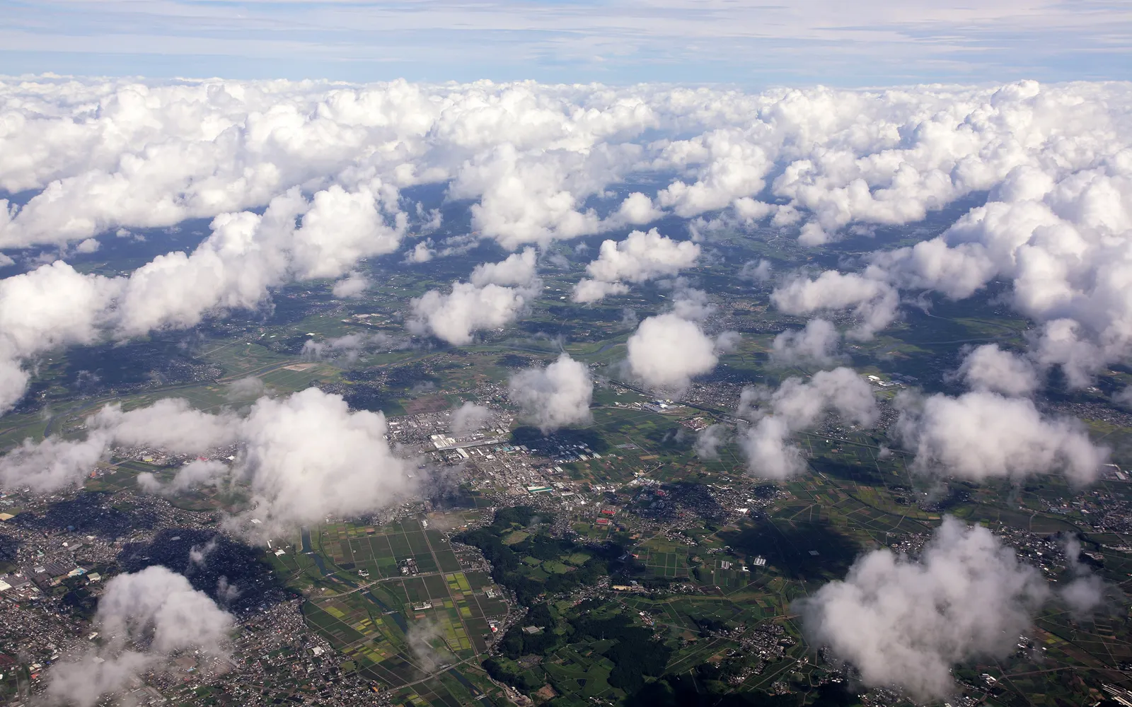 An aerial view shows fluffy white cumulus clouds scattered over a patchwork of green fields and a town.