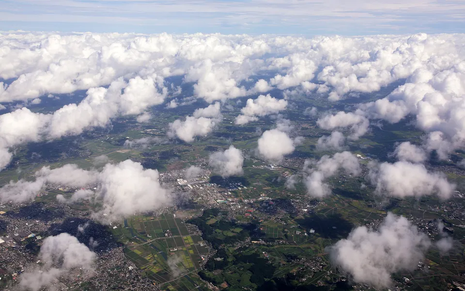 An aerial view shows fluffy white cumulus clouds scattered over a patchwork of green fields and a town.