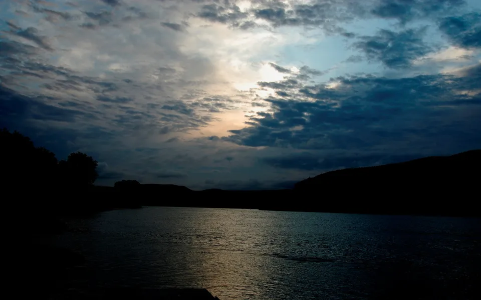 A dark, silhouetted landscape of trees and hills surrounds a calm lake under a dramatic, cloudy sky with the sun peeking through.