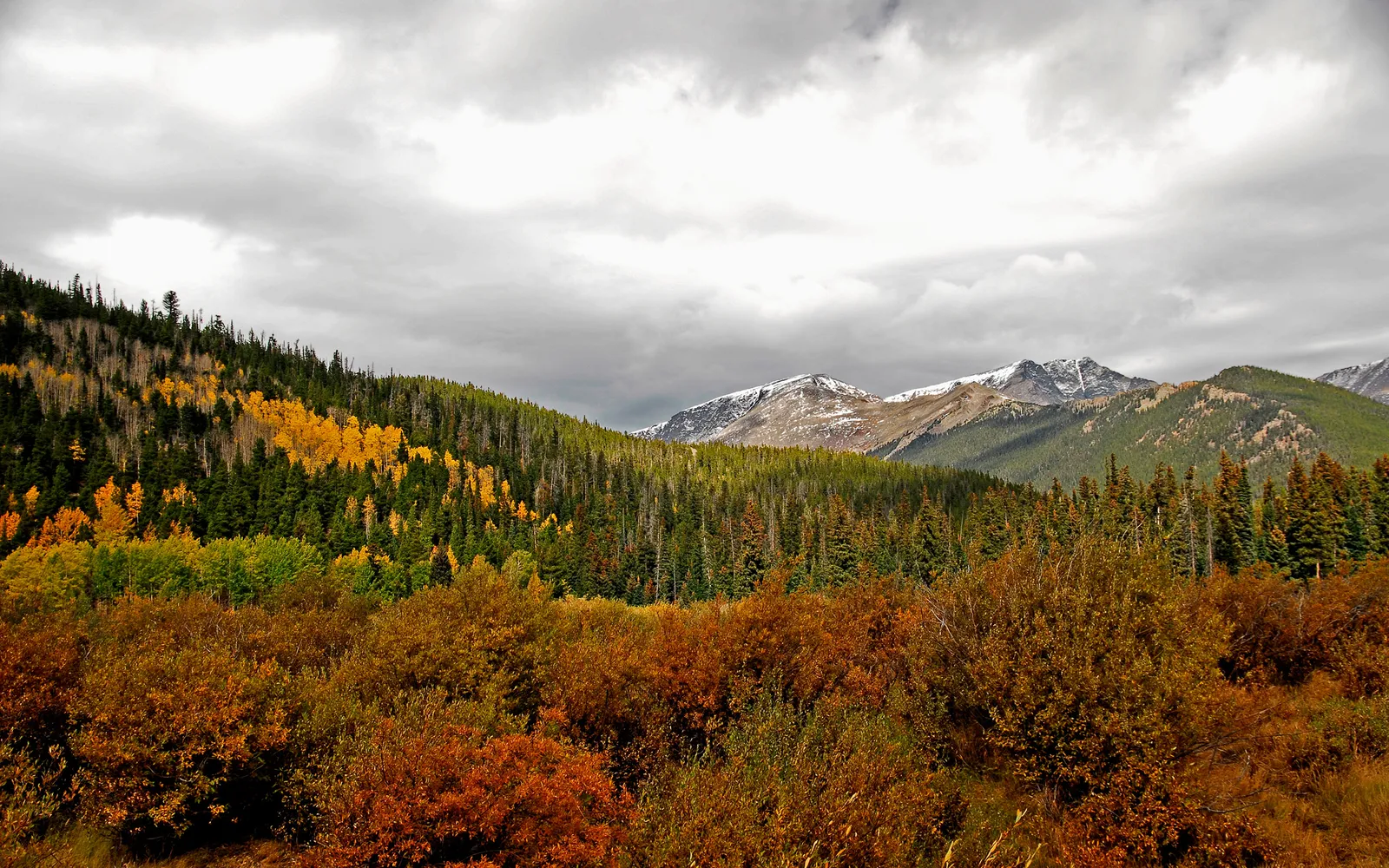 A majestic mountain range under a dramatic cloudy sky, with autumn foliage on the lower slopes.