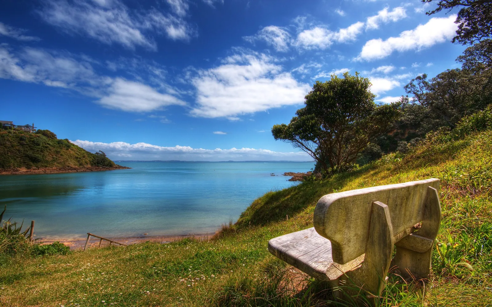 A weathered wooden bench sits on a grassy hill overlooking a calm blue bay with scattered clouds in the sky.